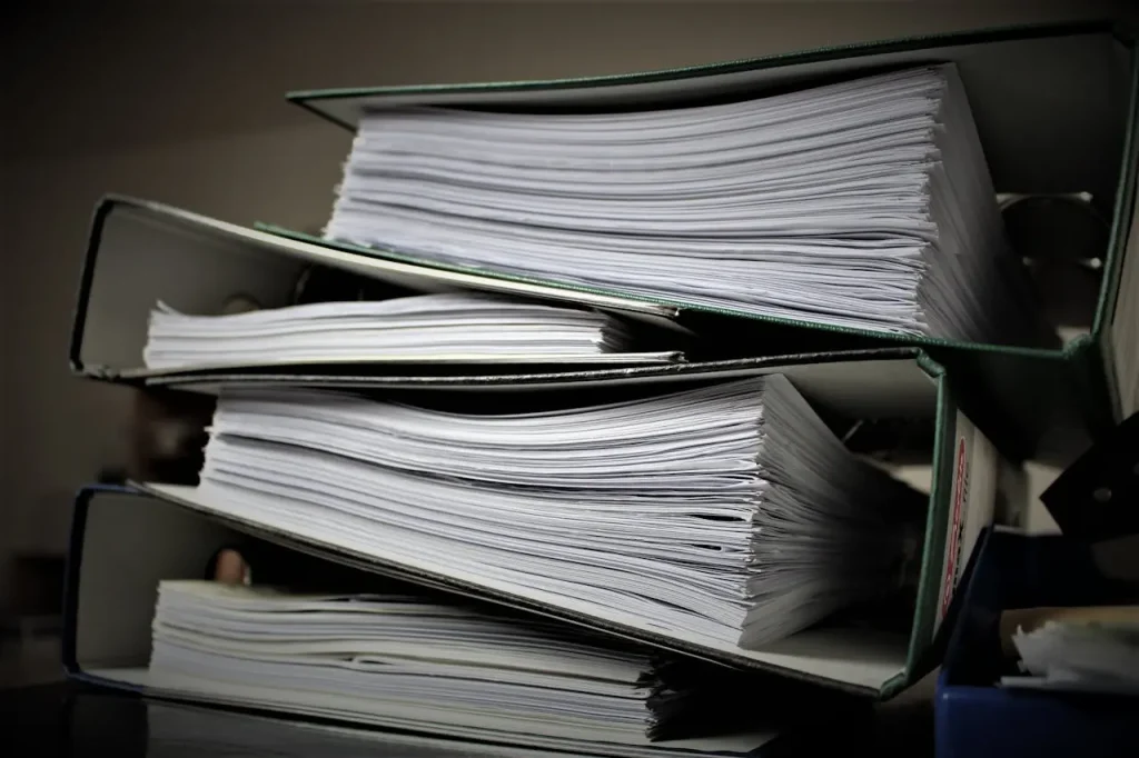 A stack of binders on a desk full of paper.