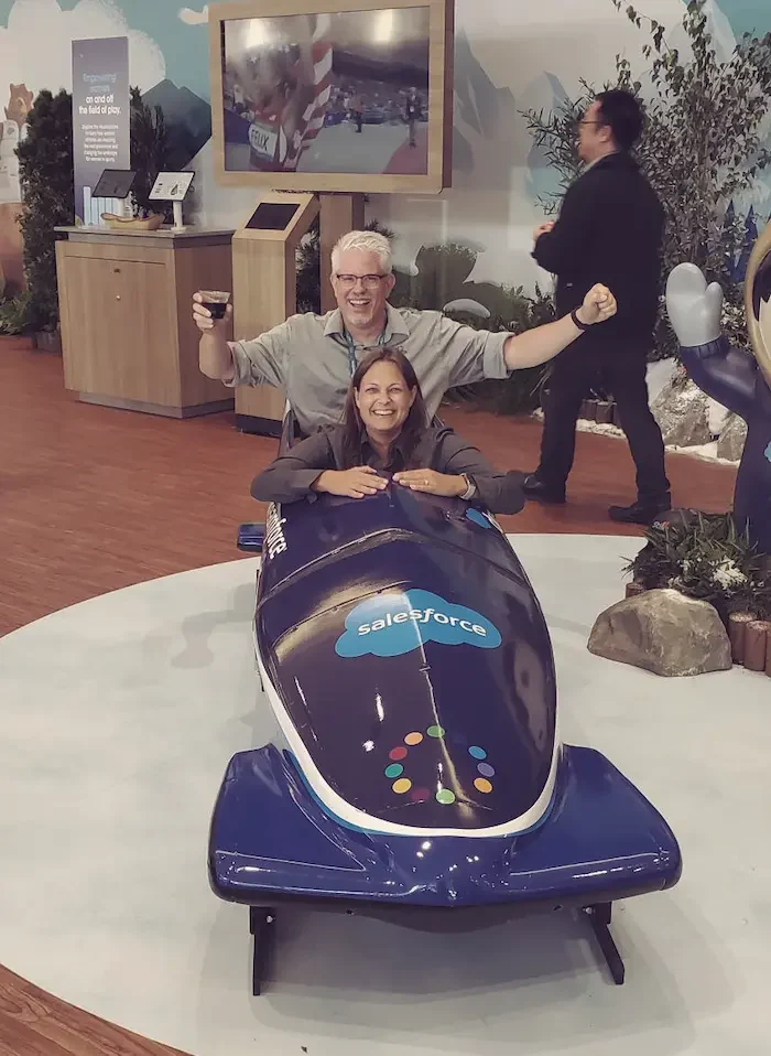 Scott Geosits and Melissa Bosch at Dreamforce having fun sitting in a blue bobsled which has the Salesforce logo printed on it.