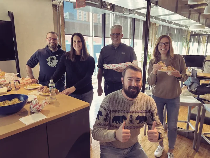 5 Trifecta employees eating lunch and smiling at the camera. One is in the foreground with two thumbs up!